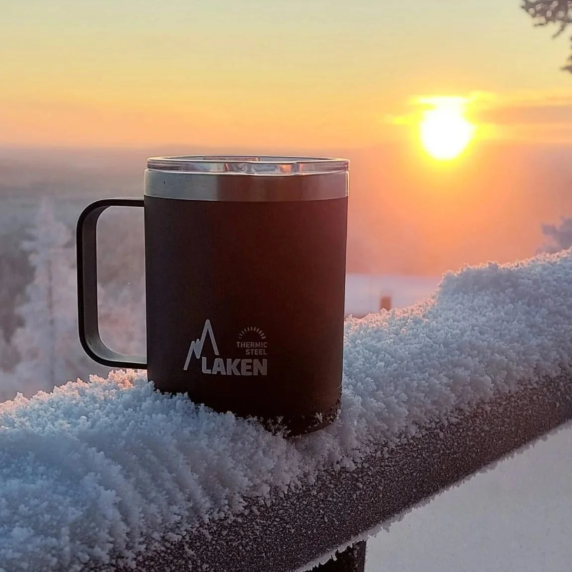 Taza térmica LAKEN apoyada en una barandilla nevada con el sol de fondo