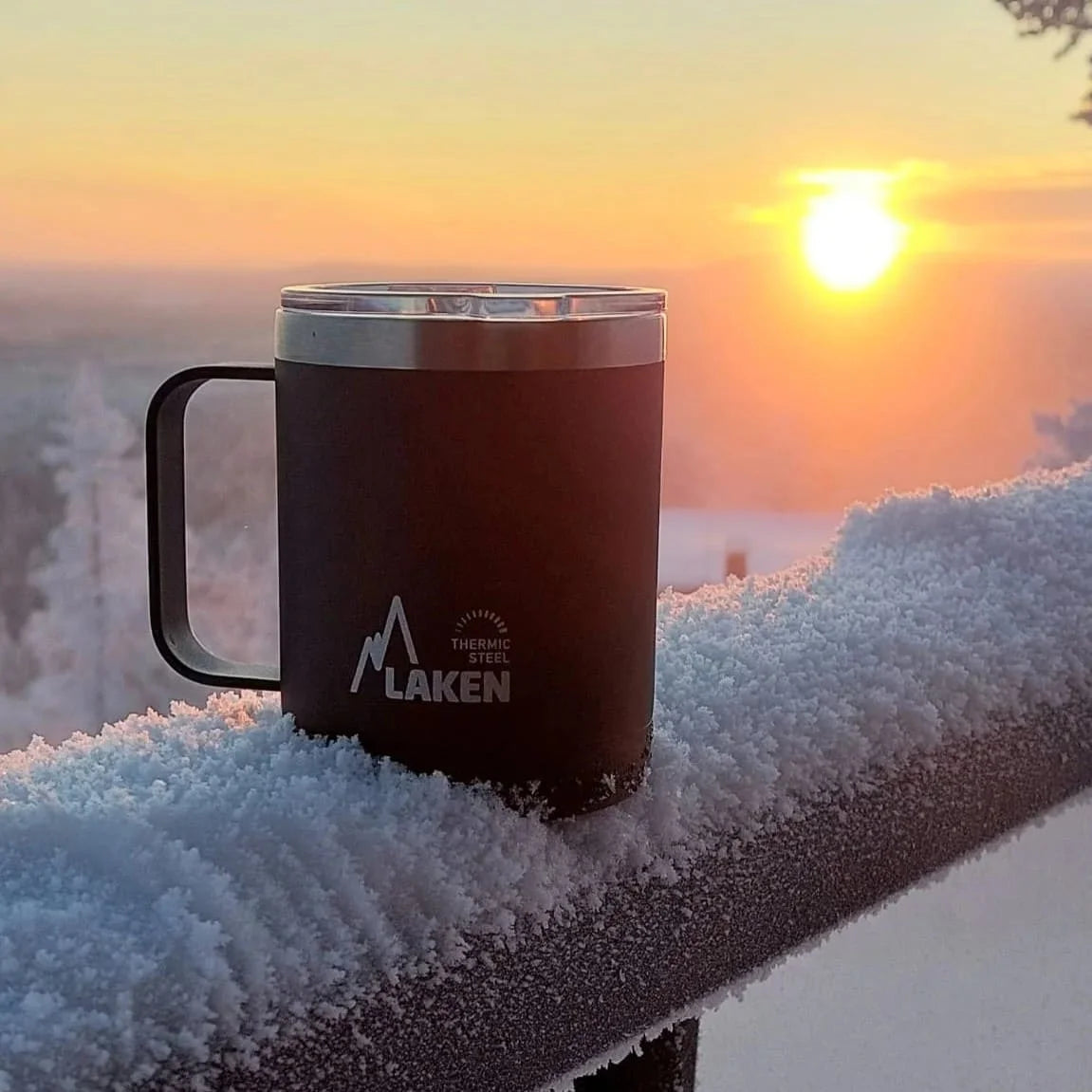 Taza térmica LAKEN apoyada en una barandilla nevada con el sol de fondo